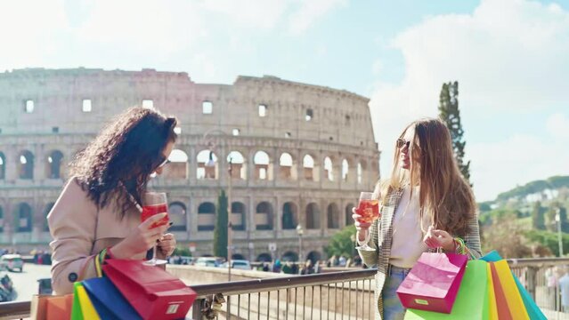 Female tourists in stylish clothes posing against ancient Colosseum background in the downtown of Rome, smiling women holding colourful shopping bags in hands and drinking refreshing aperol spritz