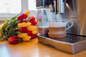 Cup of coffee with milk and tulips flowers on wooden kitchen table. Freshly brewed cappuccino espresso in coffee machine for breakfast. Stylish home interior decor. High quality photo