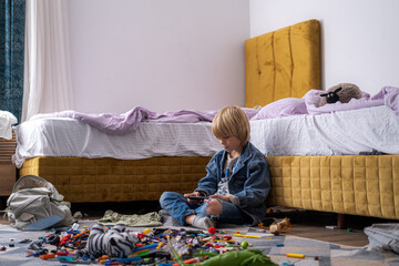 boy using phone, playing phone games in messy childrens room, kid playing among the many toys at...