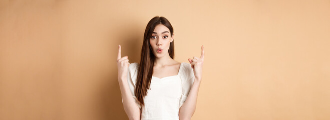 Wow look. Intrigued young woman showing interesting offer, pointing fingers up and look excited, standing on beige background