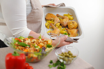 adult beautiful plump woman in apron is preparing dinner on baking sheet chicken legs quarter potatoes nearby large glass bowl with salad and nuts different ingredients she smiles