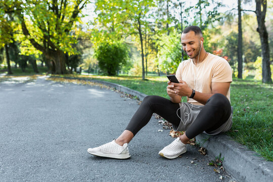 Happy and satisfied man after jogging and fitness class sitting and using smartphone, African American man in headphones listening to online podcasts, audio books and music, typing on phone.