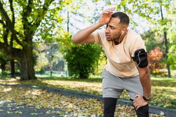 Tired man resting in park after jogging and fitness class, hispanic man using phone listening to music and podcasts in headphones breathing tired and exhausted.