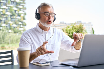 Senior older business man coach wearing headphones looking at laptop, talking having hybrid conference online remote video call, virtual distance class webinar training presentation working outdoor. - obrazy, fototapety, plakaty