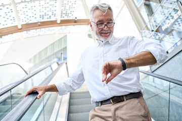Naklejka na ściany i meble Older senior business man looking at wristwatch or fitness tracker standing on escalator staircase. Mature old businessman wearing white shirt checking smart watch outside.