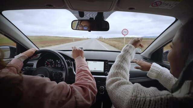 Funny Women Singing Happy While Driving Automobile. Cool Two Young Girls Dancing Lively Music In Car While Driving In The Countryside. Friends Having Fun Concept. Rear View. Slow Motion.