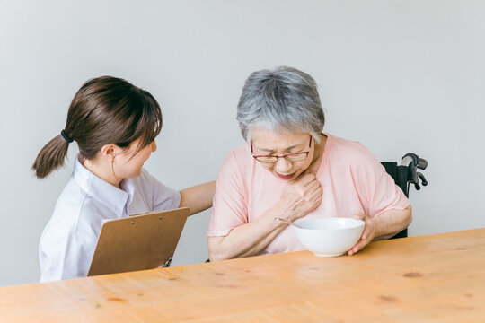 老人ホーム・家の食事で誤嚥に苦しむ高齢者女性と食事介助する管理栄養士・介護士・ヘルパーの女性
