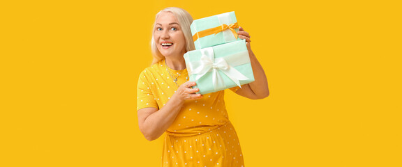 Smiling mature woman holding gift boxes on yellow background. International Women's Day celebration