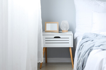 Blank frame and lamp on bedside table in light bedroom
