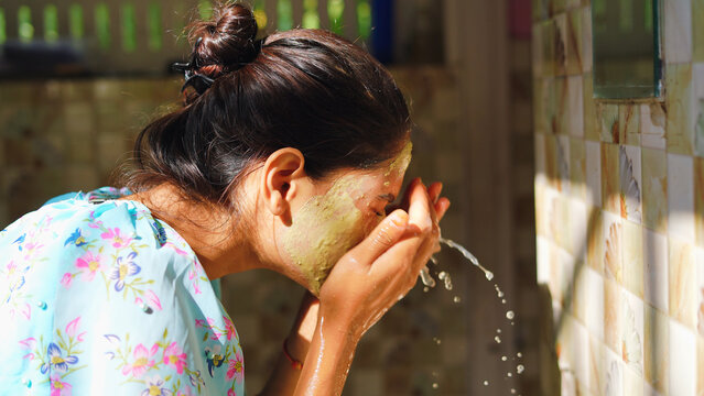 Beautiful woman is washing facial mask in bathroom after applying face mask. Girl spraying water on her face standing in front of mirror at home. mud mask or homemade multani mitti mask.