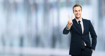 Excited businessman in black confident suit showing two fingers or victory sign gesture, over blurred interior background. Happy gesturing man. Copy space for ad slogan or text.