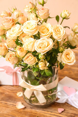 Vase with beautiful rose flowers and gifts on wooden table against color background, closeup