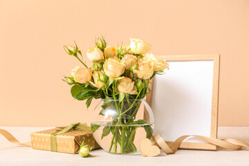 Vase with beautiful rose flowers, gift box and empty picture frame on table near color wall