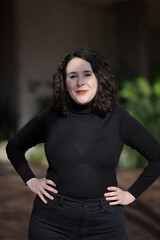 A woman with pale skin and black, wavy hair poses confidently outdoors.