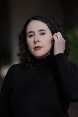 A woman with pale skin and black, wavy hair poses confidently outdoors.