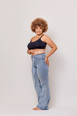 Vertical shot of body positive adult woman with curly hair holds measurable tape, looks satisfied with results with lost kilos, wears jeans and lingerie bra, poses over white wall. Concept of body