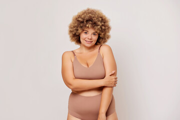 Horizontal shot of overweight adult woman with curly hair hug herself, feels confident and self loved, poses in beige lingerie over white background. Self acceptance and body positivity concept