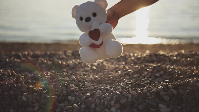 Man's hand puts white teddy bear with heart sign as gift on pebble beach in rays of setting sun on sunset. Valentine's day, Women's day, Mother's Day concepte