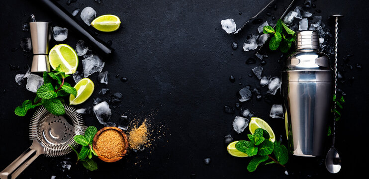 Mojito or caipirinha cocktail and mocktail preparation ingredients: mint, sugar, lime, ice and steel bar tools: shaker, jigger, strainer, muddler on dark background, top view banner
