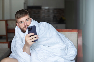 Young lazy sleepy serious man is using his smartphone, cell mobile phone at home in bedroom or living room covered in blanket, looking at screen, reading message or typing