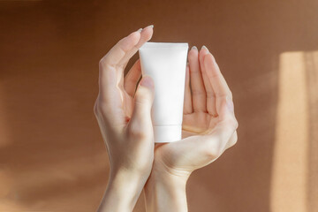 Woman's hands hold plastic container for cosmetic products. Unmarked white tube for cream, gel, lotion, toothpaste on brown background. Concept of skin care, mockup, branding