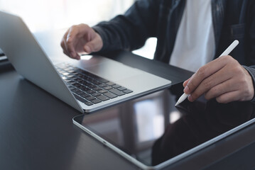 Close up of man using stylus pen signing e-document on digital tablet and working on laptop computer at home office, online working, freelancer at work, telecommuting, e-signing concept