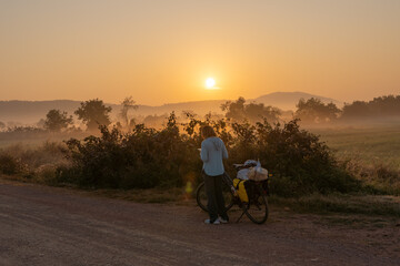Obraz premium Woman traveling on a bicycle loaded with bags on the side of a dirt road at sunrise in Cambodian countryside