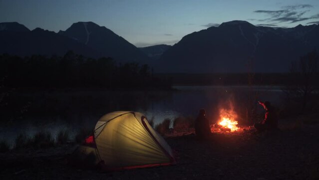 Two tourists sitting at tent with campfire and looking at the mountain. Camping