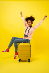 Portrait of young attractive traveling african american woman curly hair with headphone, baggage and retro camera in studio on yellow background.
