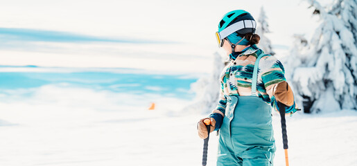 Fototapeta na wymiar Woman in skiing clothes with helmet and ski googles on her head with ski sticks. Winter weather on the slopes. On top of a mountain and enjoying view. Alpine skier. Winter sport