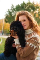 Young woman hugging her black puppy poodle dog in a park in autumn.