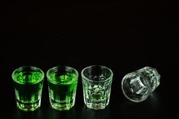 Shot glasses lined up against black background filled with emerald green spirit cocktail to be...