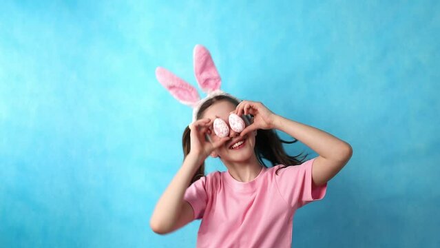 Cute teenage girl with Easter bunny ears on a blue background, keeps Easter eggs near her eyes