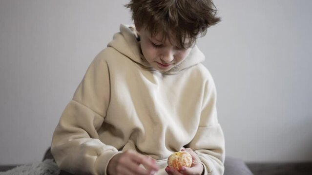 Calm Caucasian teenager peeling tangerine. Boy slowly peels the fruit and breaks it into pieces.