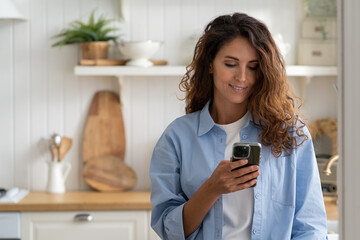 Positive smiling woman in casual clothes stands with phone in hand in kitchen in house with light interior. Cheerful optimistic girl with long hair chatting in mobile applications or typing SMS 