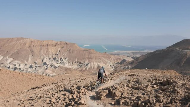 Mountain Biking Downhill steep narrow rocky ridges with scenery in the wild alone near the Dead Sea lake and Arad Mountains in the Negev Desert, Israel with bright blue sky Aerial Active Track Follow 