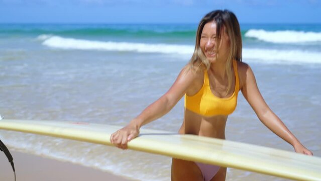4K Asian woman surfer in bikini holding surfboard walking to the ocean on tropical beach in sunny day. Attractive girl enjoy outdoor activity lifestyle water sport training surfing on summer vacation.