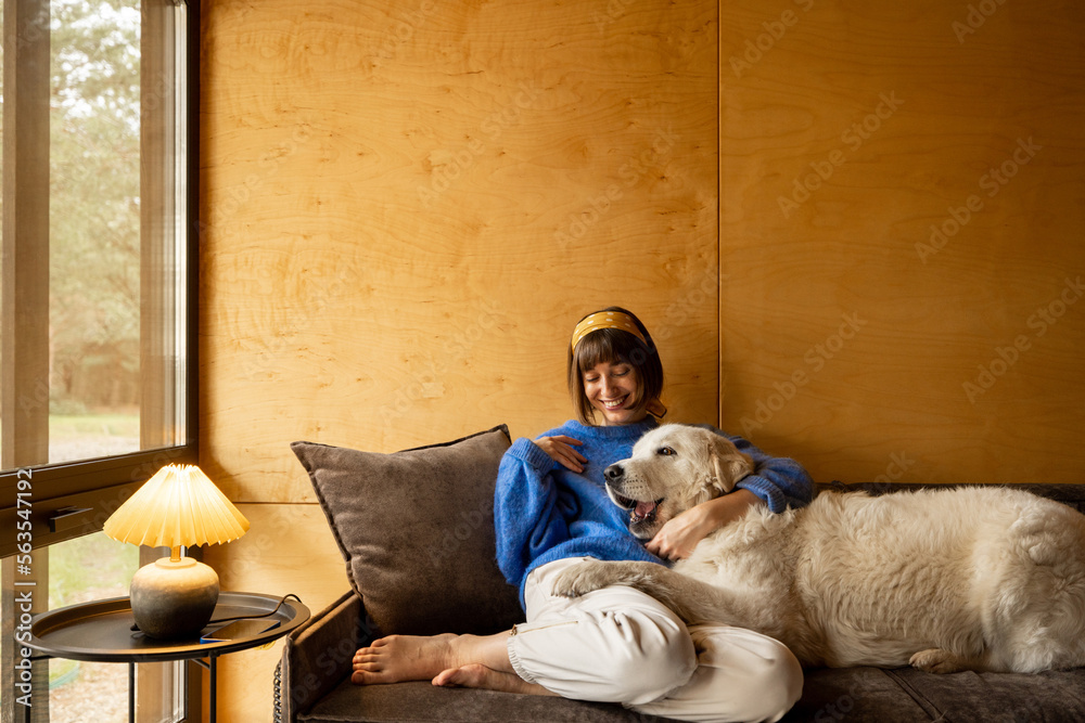 Wall mural Woman cares her dog while sitting on a couch by the window, spending leisure time together in wooden house on nature