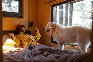 Young woman cares her huge adorable white dog while lying in bed in tiny bedroom of wooden cabin on...