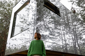 Woman near an invisible house with mirrored walls in pine forest. Connection with nature and sustainability concept. Rest in tiny cabins on nature