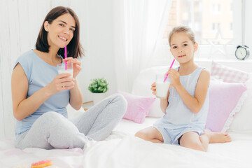 Obraz na płótnie Canvas Happy brunette young mum and her pretty daughter sit on bed, drink milk shake and eat delicious doughnuts, enjoy calm domestic atmosphere in bedroom. People, leisure, bedding, motherhood concept