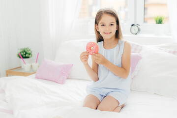 Obraz na płótnie Canvas Lovely small girl holds delicious doughnut in hands, going to have breakfast, poses on bed in cozy white bedroom, looks at camera with positive expression. Children, dessert, bed time concept
