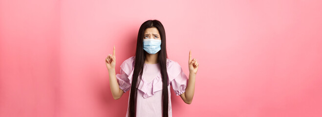 Coronavirus, quarantine and lifestyle concept. Disappointed sad asian girl pointing fingers up,...