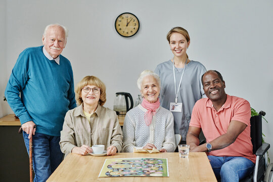 Portrait of senior people with caregiver smiling at camera while playing board game - Powered by Adobe