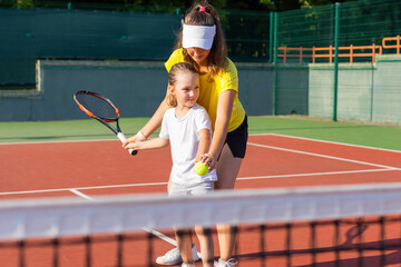 Cheerful coach in sports clothing teaching child to play tennis while both standing on tennis court.