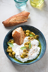 Bowl of ricotta cheese served with crusty bread, green olives and olive oil, vertical shot on a light-beige stone background