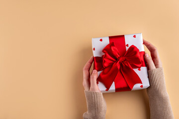 Female hands holding present with red bow and white paper in hearts on beige background. Valentine's day gift wrapping concept.