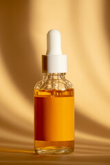 Orange serum in transparent glass bottle with white dropper lid in rays of morning sun on brown...