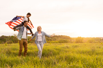 Naklejka na ściany i meble Young parents with their daughter holding American flag in countryside at sunset. Independence Day celebration