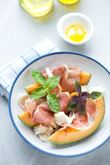 Plate with cantaloupe slices, prosciutto, torn mozzarella and basil leaves, vertical shot on a light-grey granite background